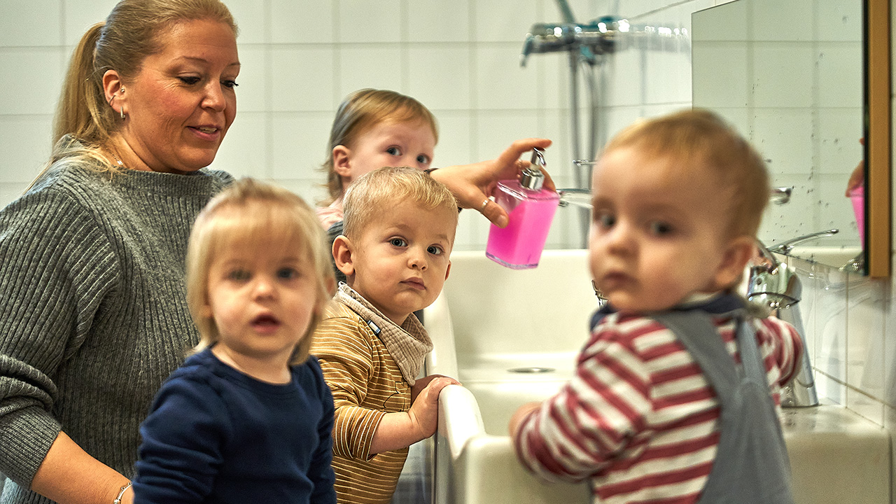 Drei kleine Kinder stehen vor einem Waschbecken. Eine Frau hinter ihnen hat einen Seifenspender in der Hand