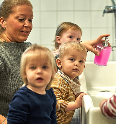 Drei kleine Kinder stehen vor einem Waschbecken. Eine Frau hinter ihnen hat einen Seifenspender in der Hand