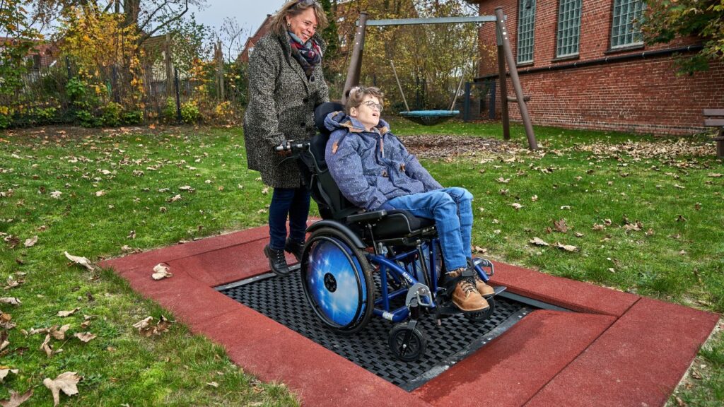 Eine Frau steht mit ihrem Rollstuhl zusammen mit einer anderen Frau auf einer Art Trampolin.