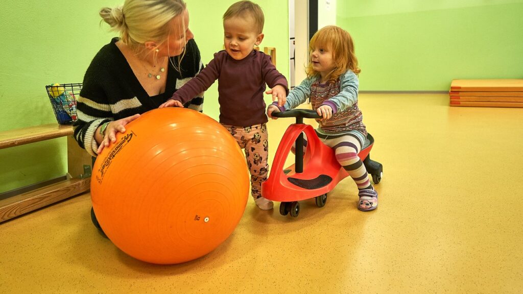 Eine Frau sitzt neben einem Kind auf einem Laufrad und ein Kind mit einem kleinen Gymnastik-Ball