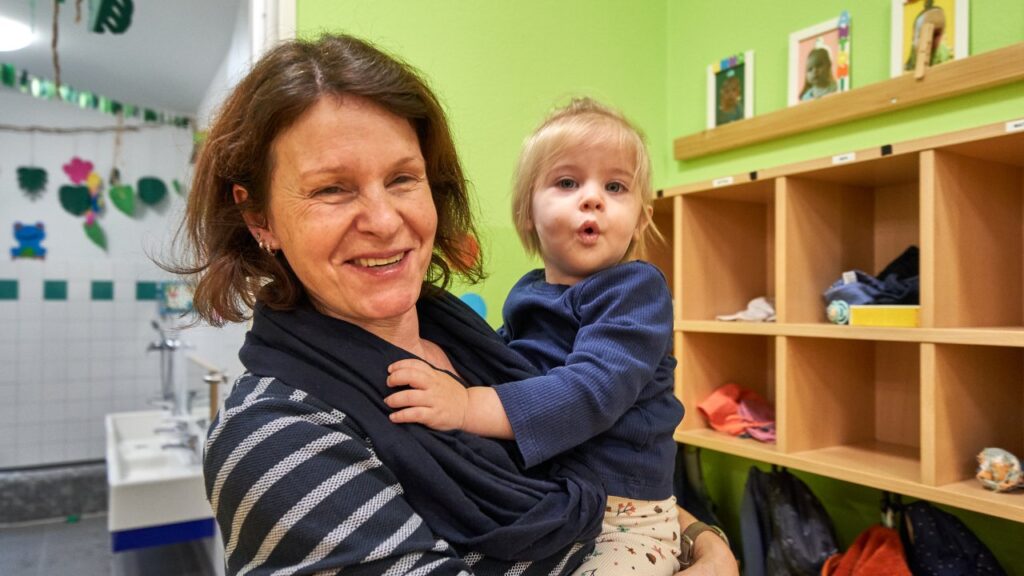 Lachende Frau hat ein Kind auf dem Arm. Beide schauen in die Kamera