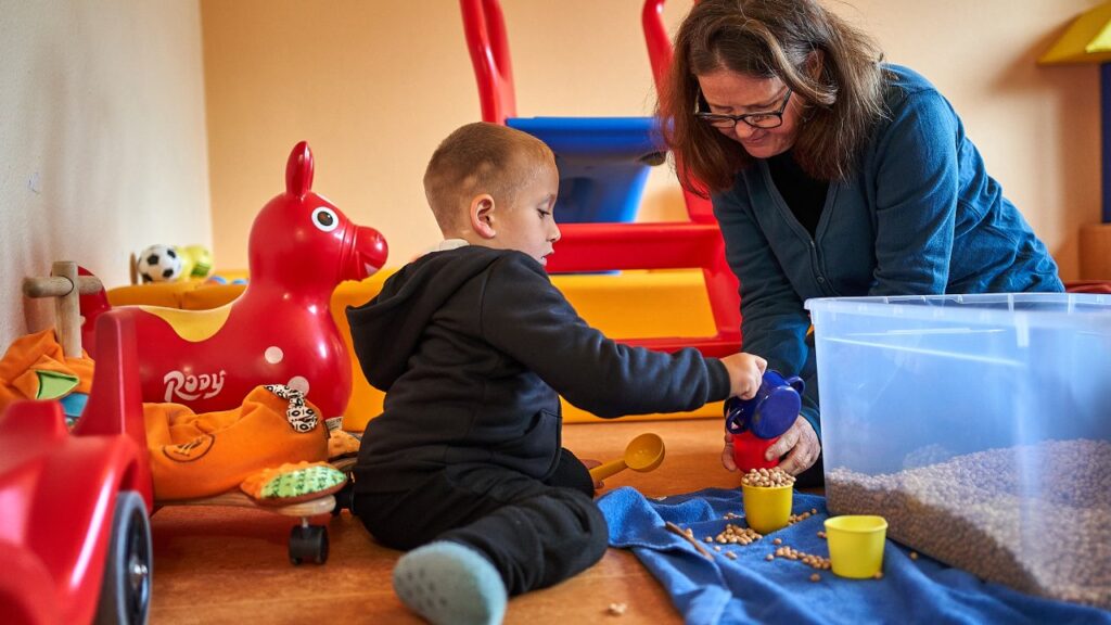 Kind füllt mit Hilfe einer Frau Linsen von einem Gefäß in ein anderes