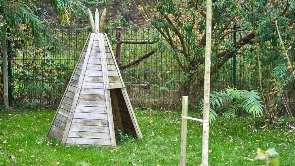 Ein Holztipi auf einer Rasenfläche. Im Hintergrund Bäume, Büsche und ein Zaun